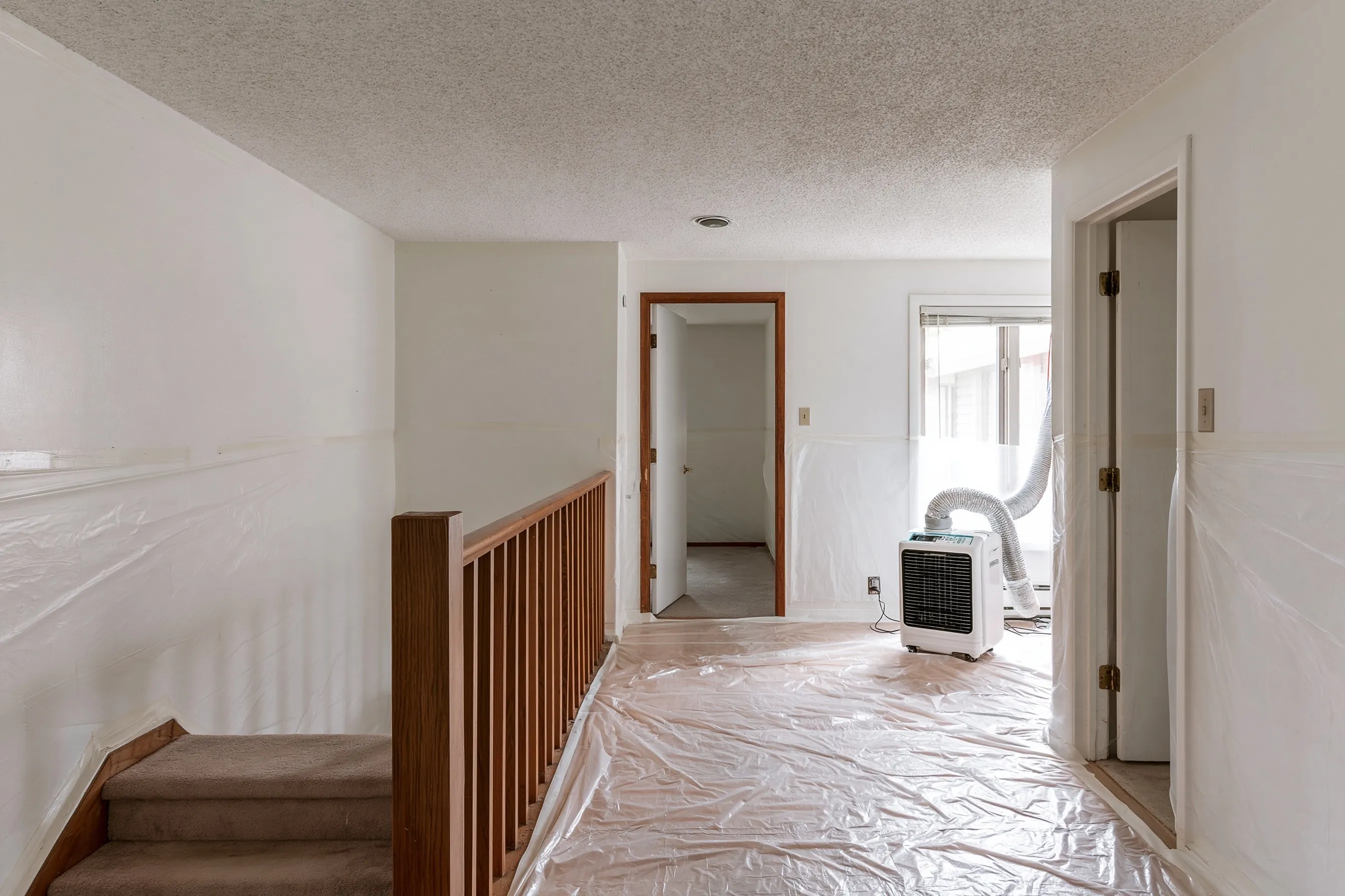 Upper floor of a house with a painted popcorn ceiling, plastic over floor and walls, a half-flight of stairs in view