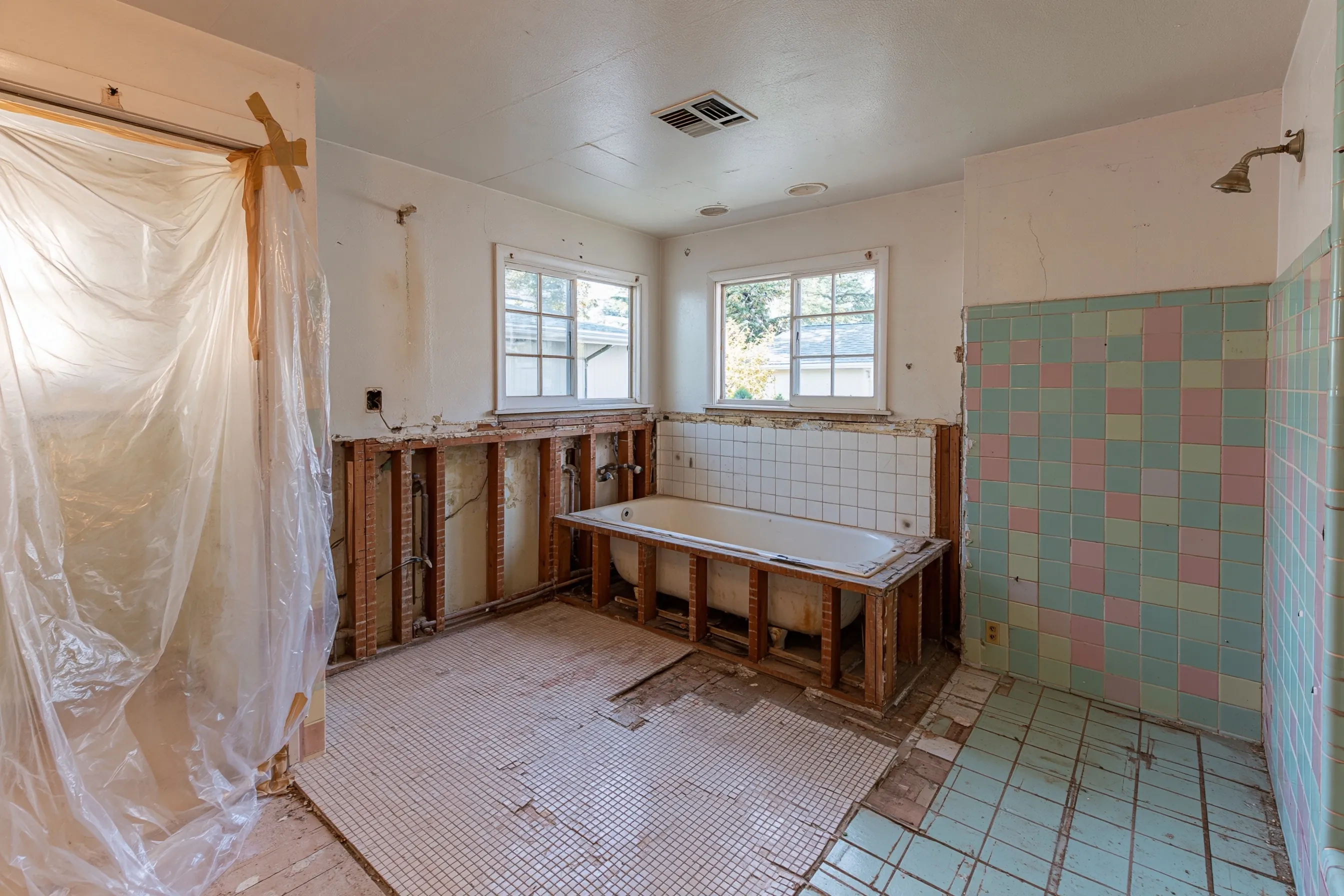 A bathroom opened for a remodel with the tub-surround wall cut out, floor tile lifted and plastic up